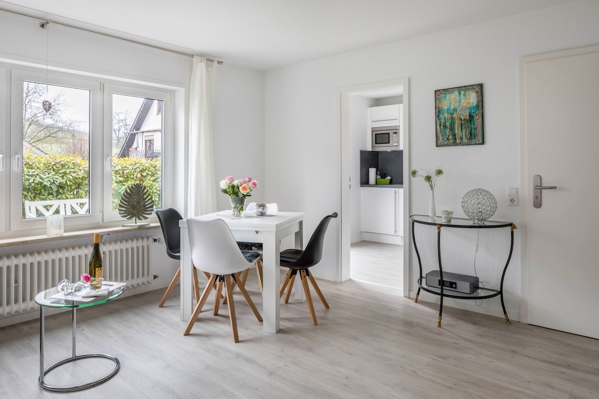 A bright living area features a white table surrounded by black and white chairs. A small glass side table holds decorative items. Natural light floods through a large window, which overlooks greenery outside. The adjoining kitchen is partially visible, showcasing modern appliances.