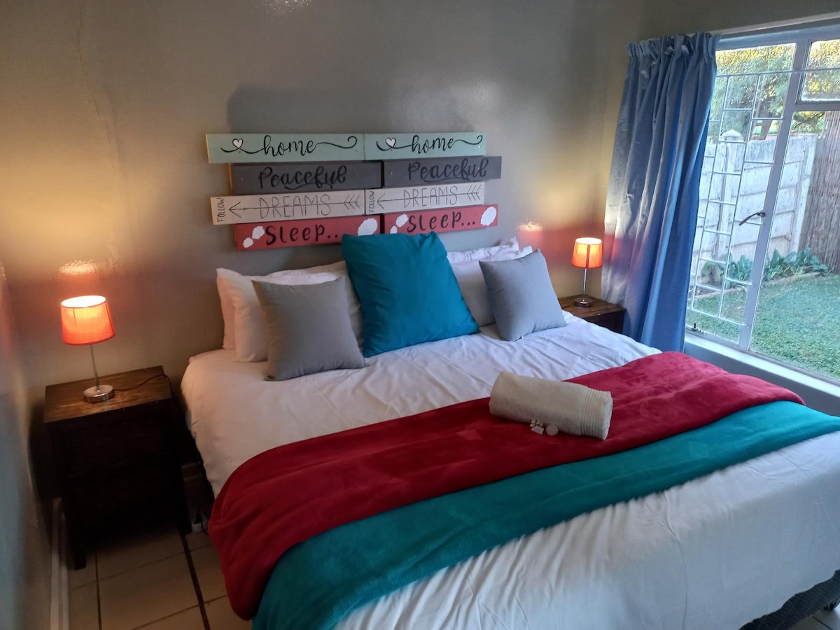 A sleeping area features a spacious bed with decorative pillows and a vibrant throw blanket. Soft bedside lamps illuminate the nightstands, while a large window with light blue curtains allows natural light to enter, revealing a glimpse of the greenery outside.