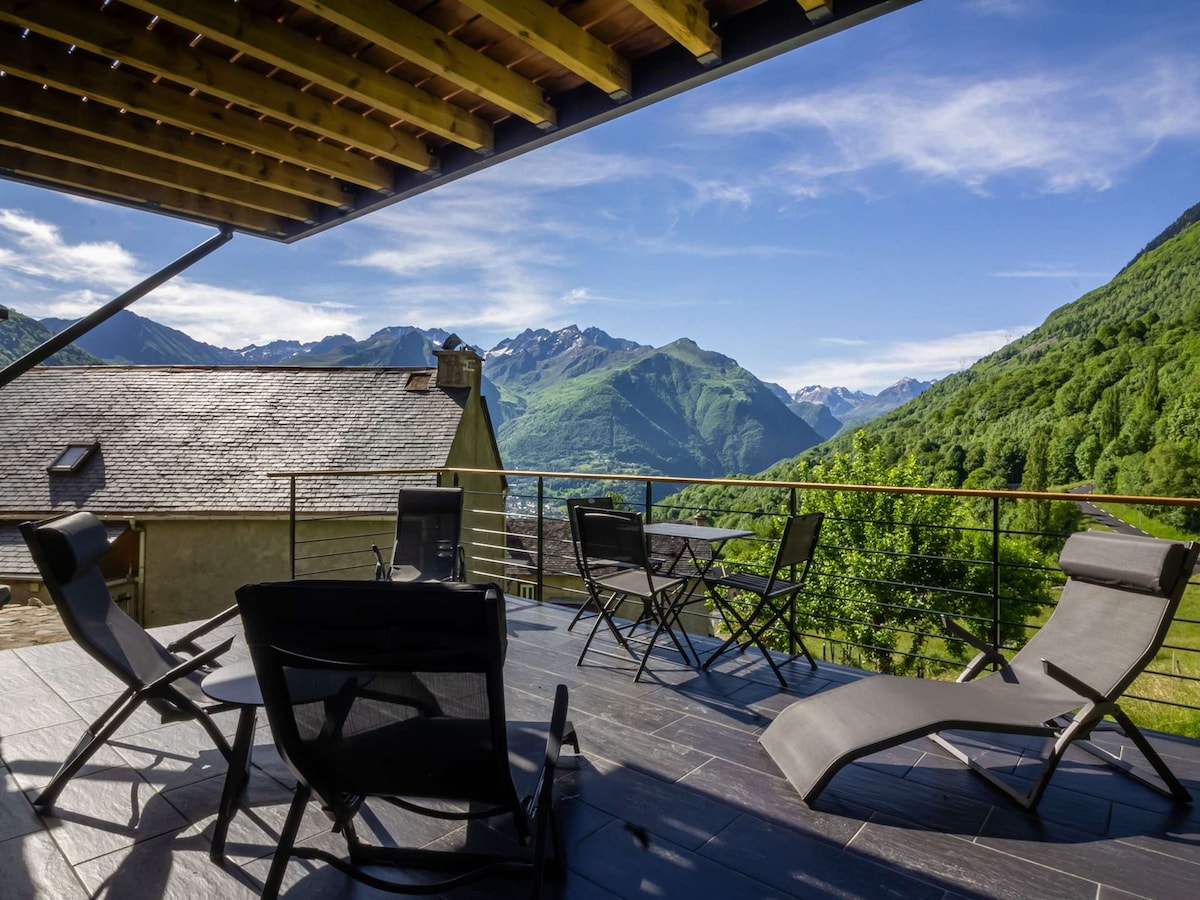 An outdoor terrace features several lounge chairs arranged for relaxation. A scenic view of green mountains and valleys is visible in the distance, with clear blue skies overhead. The terrace is partially covered, providing a shaded area for guests.