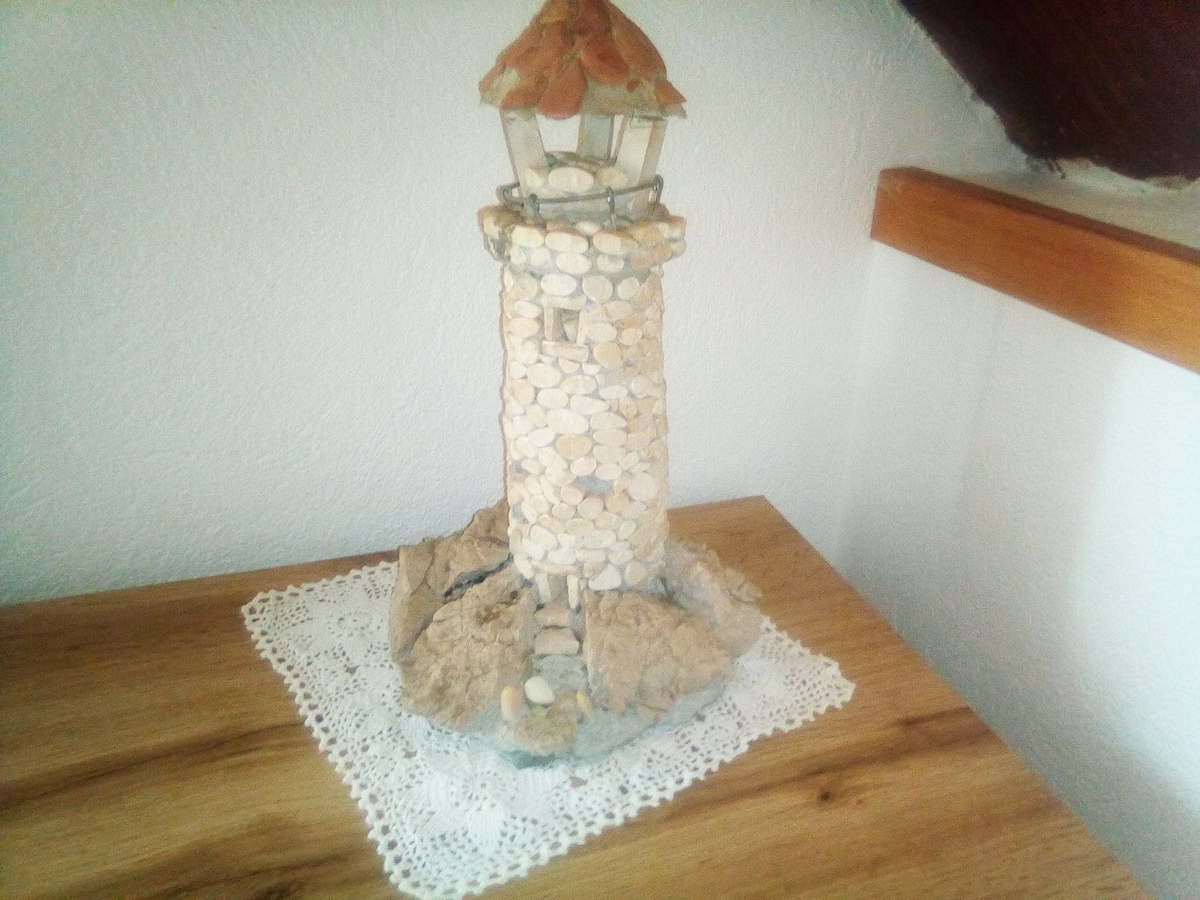 A decorative lighthouse sculpture is positioned on a wooden table. The lighthouse, crafted from pebbles and stones, features a distinct orange roof and detailed textures. A lace doily is placed beneath the sculpture, adding a touch of elegance to the natural display.