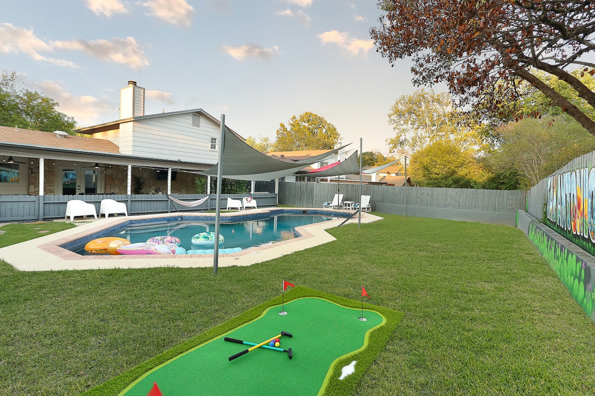 A private swimming pool is set within a spacious yard, surrounded by green grass and shaded by a large awning. Colorful pool floats are arranged in the water. Nearby, a small putting green is visible, inviting leisure activities.