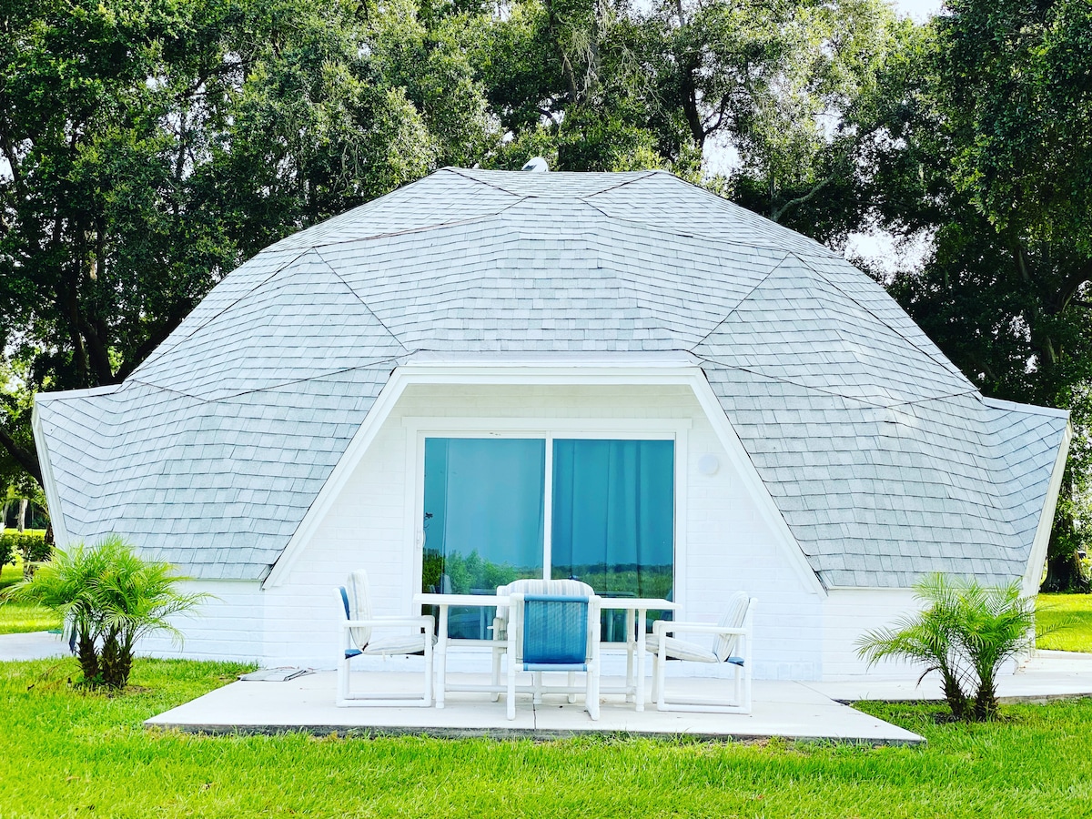 A unique, modern structure features a multi-faceted roof and large glass windows that reflect the surrounding greenery. An outdoor patio is furnished with a table and chairs, providing a serene spot to enjoy the natural landscape. Lush grass surrounds the building.