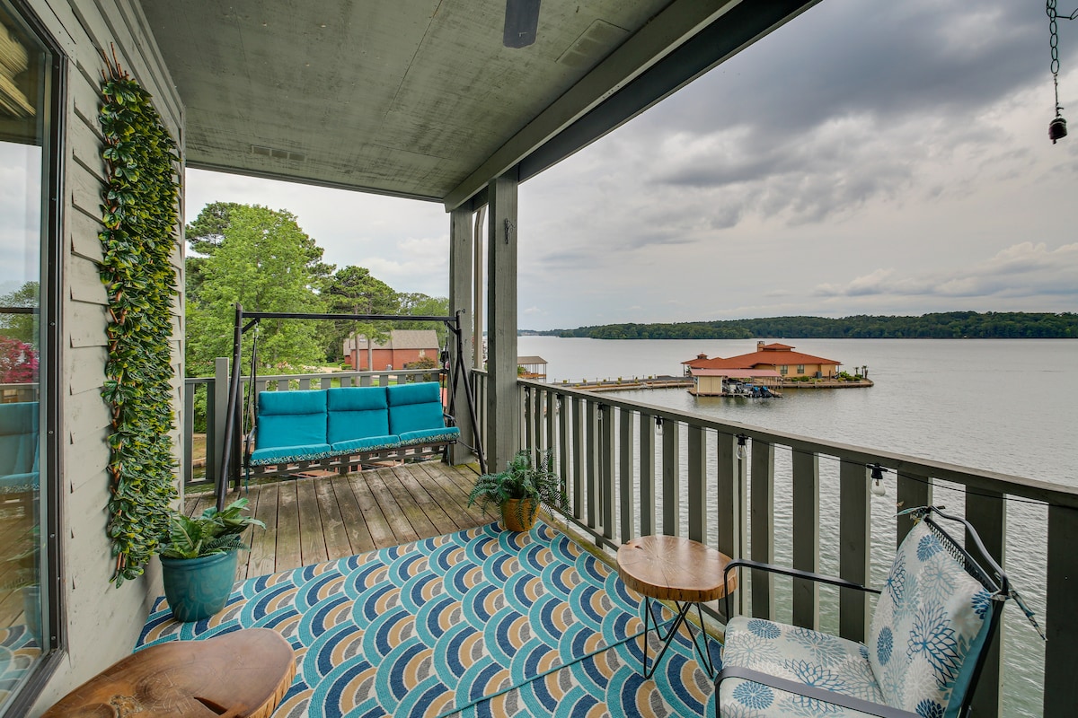A wraparound balcony offers panoramic views of Lake Hamilton, featuring a bench swing and cushioned seating. Outdoor tables are complemented by a patterned area rug, and potted plants add touches of greenery, inviting relaxation in a serene lakeside setting.