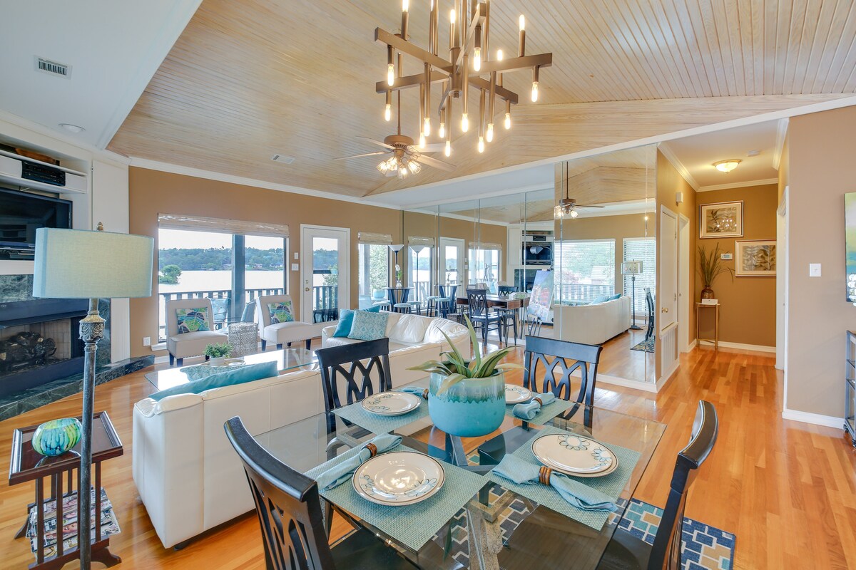 An open-concept living area is highlighted by natural wood beams and large windows offering scenic lake views. A dining table is set for four, complemented by modern decor. A cozy seating arrangement surrounds a gas fireplace, creating a welcoming space for gatherings.
