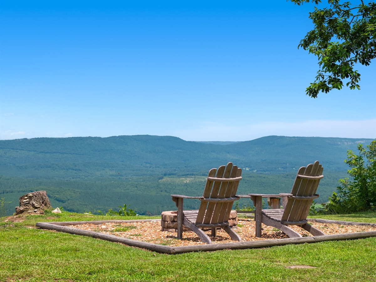 Two adirondack chairs are positioned on a gravel surface, facing a panoramic view of rolling hills and distant mountains under a clear blue sky. A small fire pit is located between the chairs, suggesting a space for gatherings and contemplation.