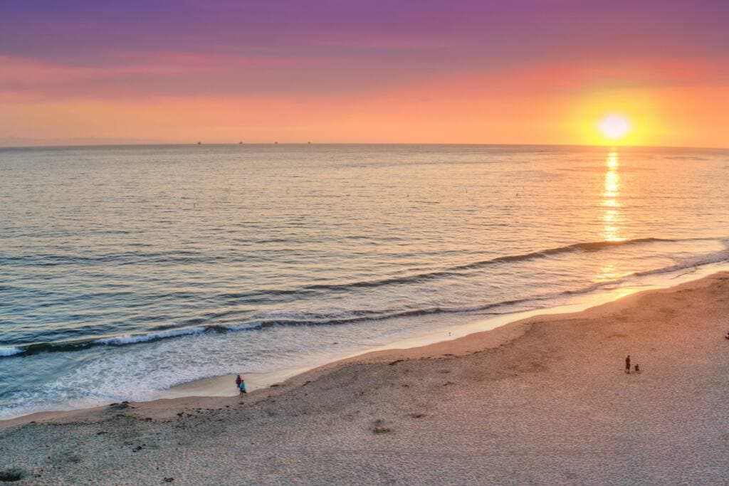 A serene beach view captures gentle waves lapping at the shoreline during sunset. The sky displays soft hues of purple and orange as the sun dips toward the horizon, setting a tranquil mood for the coastal landscape.