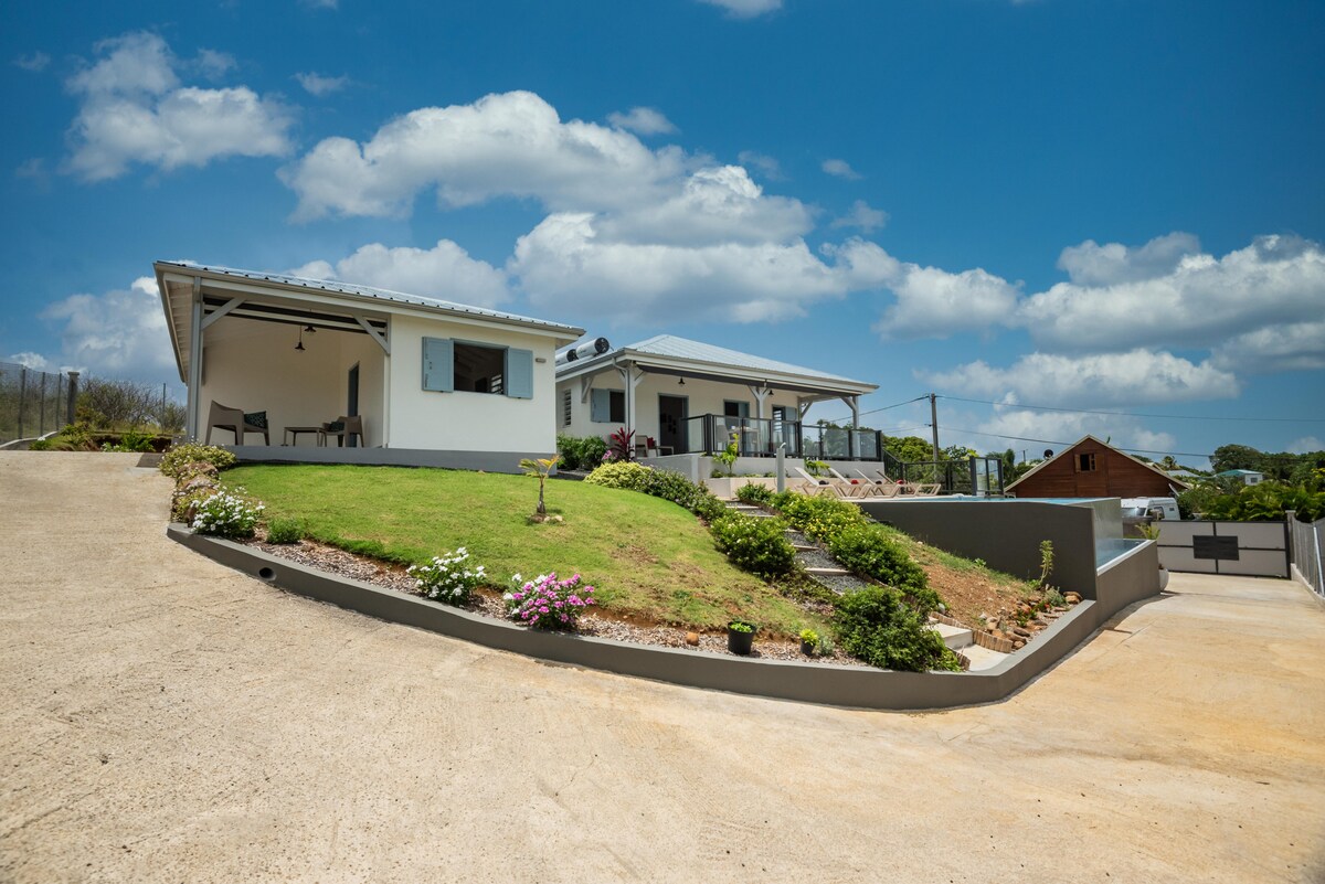 The exterior of Maison Sugar Cane is showcased with a well-maintained lawn and flowering plants. The structure features a charming entrance with a covered porch, and two distinct units are visible, set against a clear blue sky with scattered clouds.