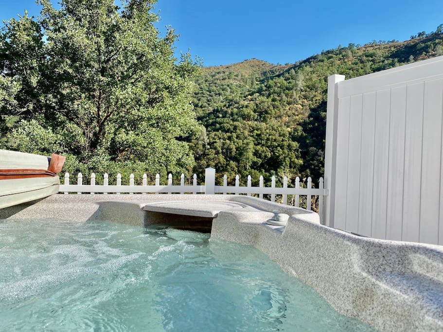 A private hot tub is positioned adjacent to a white picket fence, overlooking lush green hills. The water is clear with gentle bubbling, inviting relaxation while enjoying the surrounding nature.