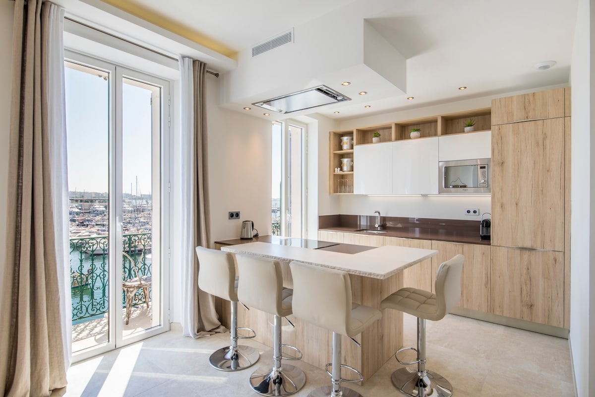 A modern kitchen features a large island with four bar stools, surrounded by light wood cabinetry and sleek white countertops. Natural light floods through the large windows, offering a view of the marina.
