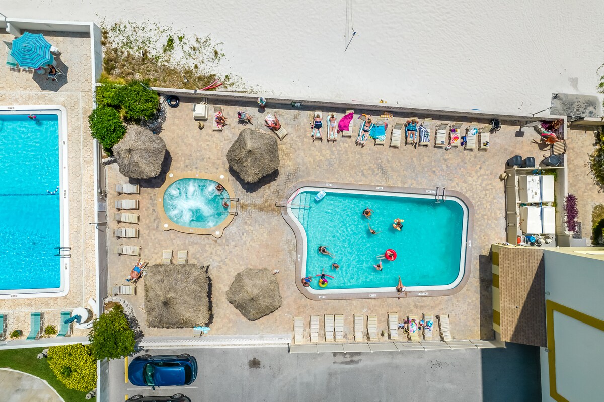 An aerial view showcases a pool area surrounded by sandy beach. Two pools are visible, one larger with a hot tub nearby. Sun loungers and umbrellas are arranged around the pools, with various guests enjoying the amenities and shade provided by thatched-roof cabanas.