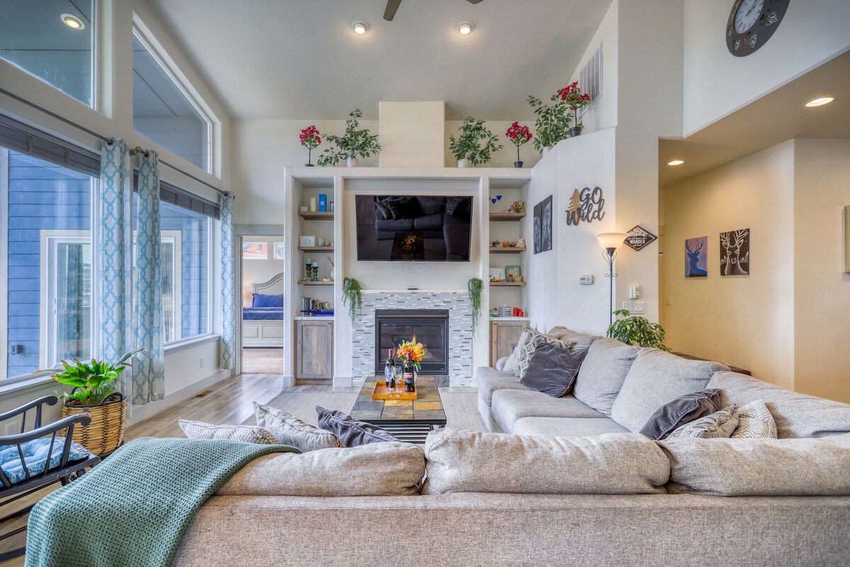 A spacious living area features a large sectional sofa adorned with a cozy throw, positioned in front of a modern stone fireplace. Floral decorations and shelves filled with decorative items enhance the ambiance, while large windows allow ample natural light to filter in.