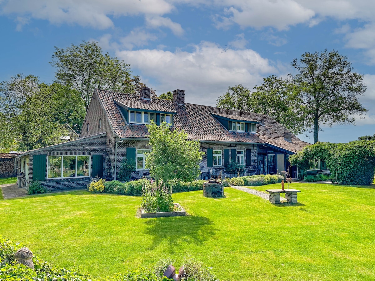 The spacious exterior of a traditional farmhouse is surrounded by a well-maintained green lawn. Dense trees and bushes create a natural border, while the building features a combination of windows and doors framed with contrasting shutters.