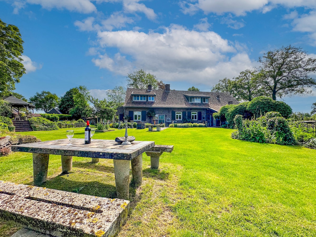 A spacious garden features a large stone table with two benches. Lush green grass surrounds the area, while a charming farmhouse with multiple windows provides a scenic backdrop. Mature trees and well-maintained hedges enhance the outdoor setting.