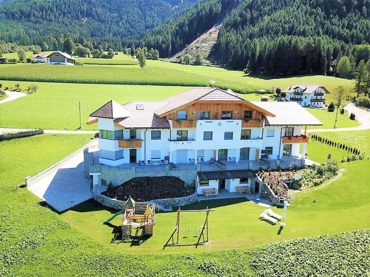 The modern apartment building is positioned amidst lush greenery and gentle hills. It features multiple balconies and large windows, maximizing natural light. A playground with swings is visible in the yard, alongside well-maintained gardens inviting outdoor activities.