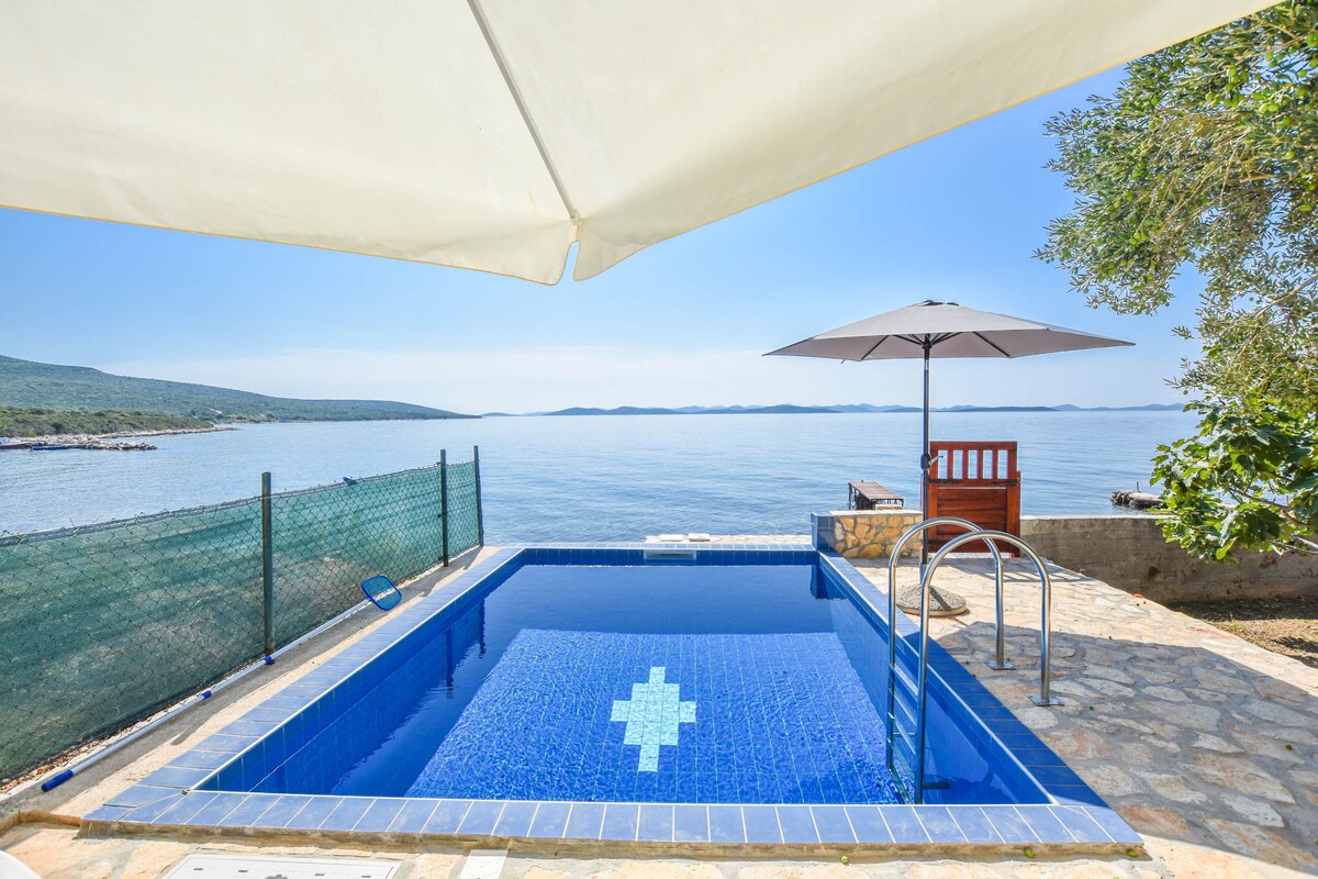 A sunlit outdoor pool is surrounded by a stone terrace, offering views of the calm sea and distant islands. A large umbrella provides shade beside the pool, while a ladder allows easy access to the water. Nearby, a wooden seating area adds to the tranquil setting.