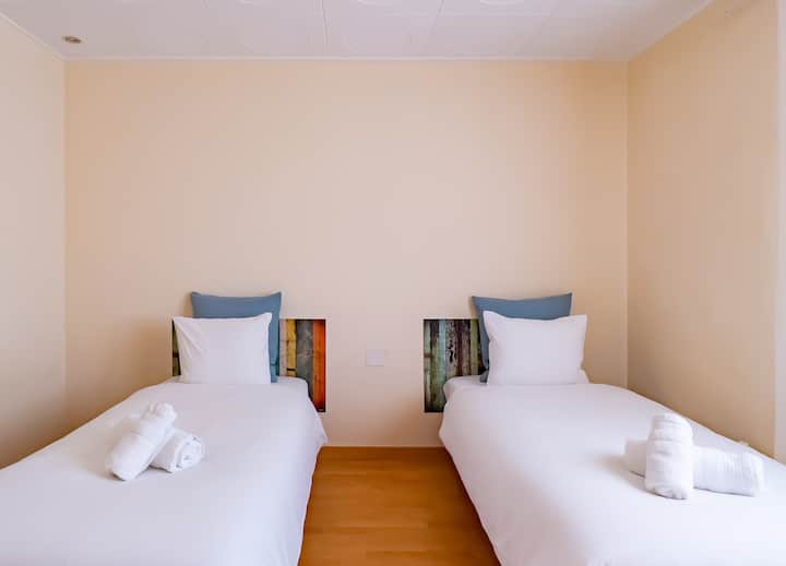 Un luminoso y acogedor dormitorio doble con dos camas individuales vestidas con sábanas blancas frescas, cada una con una cabecera de madera colorida y única. Las suaves paredes beige y la decoración minimalista crean un ambiente acogedor pero moderno.
