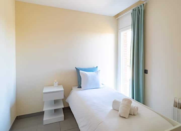 Una habitación individual luminosa y acogedora con una cama cuidadosamente hecha con sábanas limpias, una mesita de noche elegante y una gran ventana que permite que la luz natural llene el espacio. La suave decoración azul y blanco crea un ambiente tranquilo y relajante.