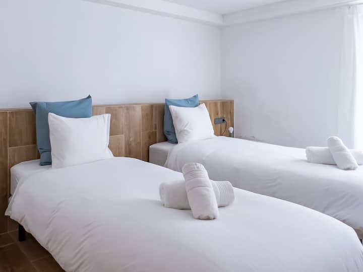 Un dormitorio doble luminoso y elegante con dos camas individuales con sábanas blancas nuevas, complementadas con cabeceras de madera y almohadas azules suaves. La luz natural entra por la ventana, creando un ambiente sereno y cómodo.