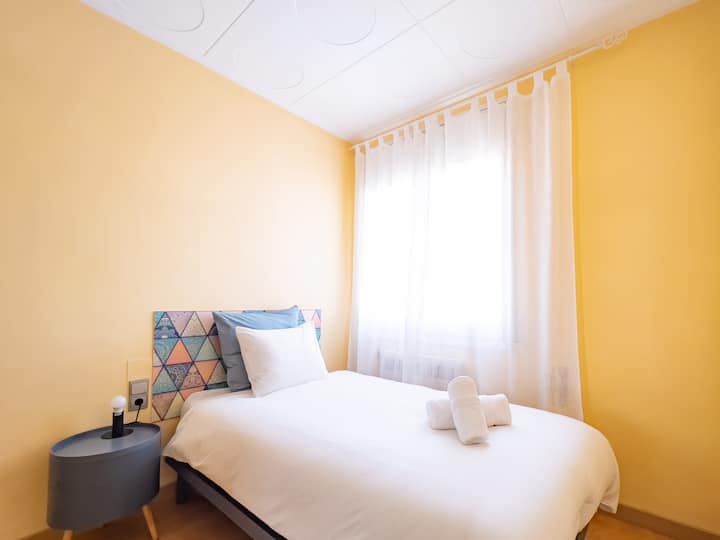 Una acogedora habitación individual con una cómoda cama, sábanas blancas nuevas y una cabecera colorida de diseño único. Las cálidas paredes amarillas y la luz natural de la ventana crean un espacio luminoso y acogedor.