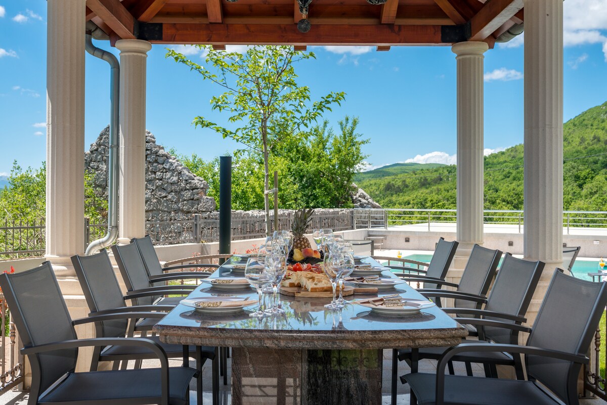 An outdoor dining area features a large stone table surrounded by several chairs, set against a backdrop of lush greenery and clear blue skies. Table settings are arranged with plates and glasses, ready for a meal. A private pool is visible in the distance.