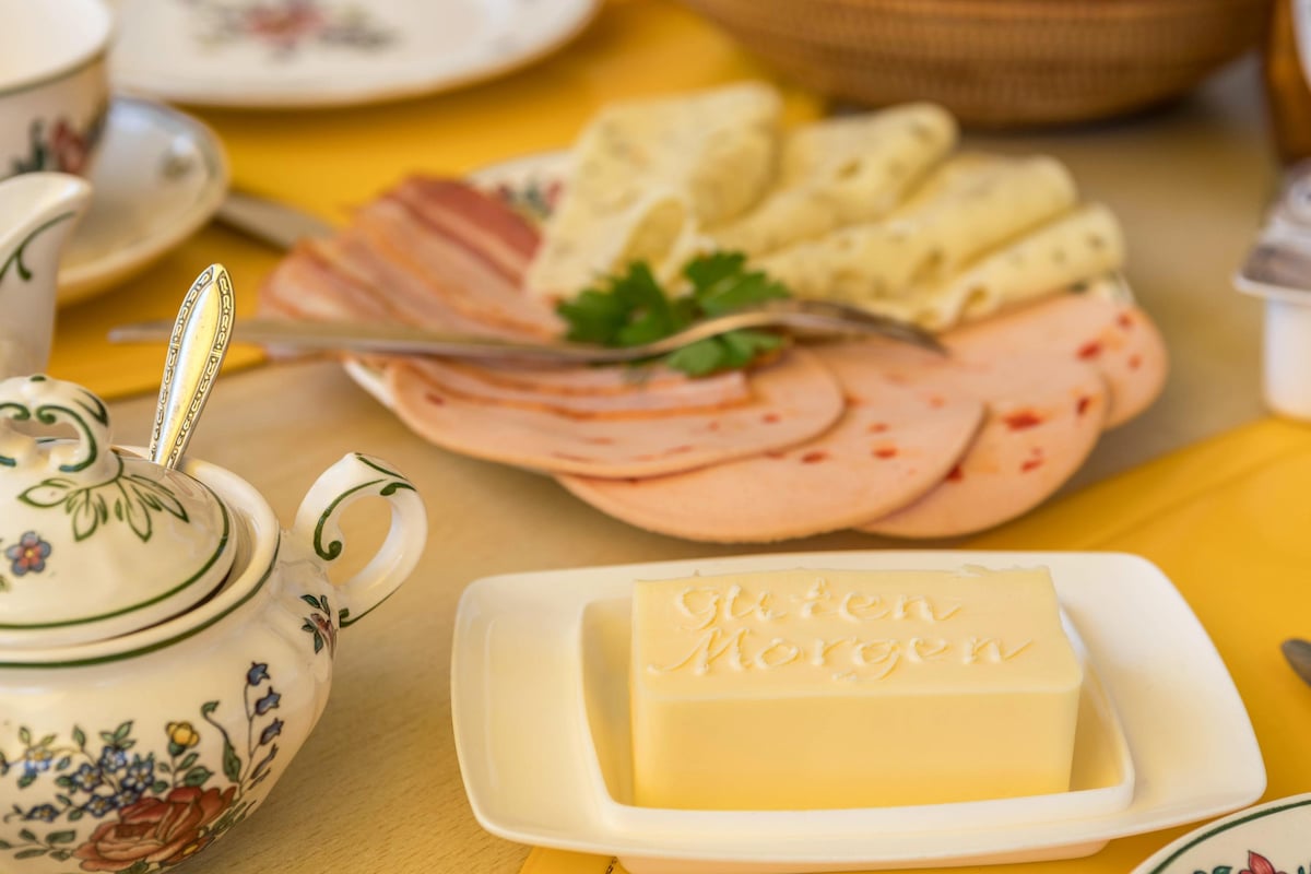 A breakfast spread is presented, featuring a variety of sliced meats and cheeses arranged on a decorative plate. A butter block with 'Guten Morgen' etched into it sits nearby, alongside delicate porcelain serving pieces. The table is adorned with a bright yellow tablecloth.