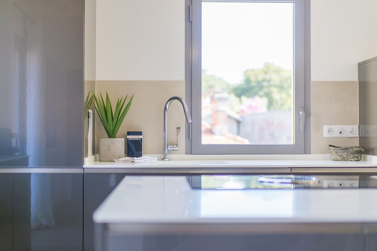 A modern kitchen showcases sleek cabinetry and a central island with a flat surface. Natural light enters through a large window, illuminating the space. A small potted plant sits on the countertop, complementing the minimalist design.