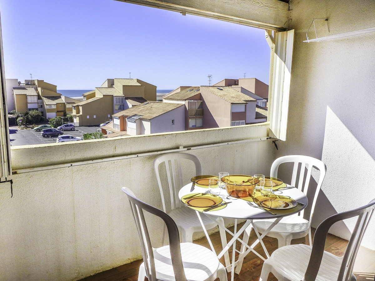 A dining table is set on the loggia, featuring white chairs and a circular table adorned with yellow plates and glassware. The scene showcases a view of neighboring buildings and the sea beyond, with bright light enhancing the spacious ambiance.