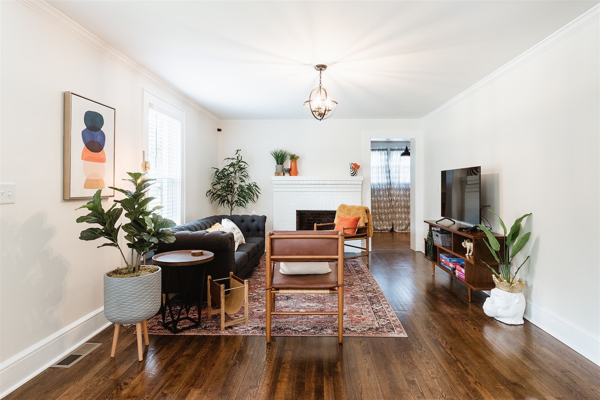 The living room features a comfortable black sofa paired with a mid-century wooden chair. A circular coffee table sits on a traditional area rug, and greenery is presented in a decorative planter. Natural light enters through the window, illuminating the space's neutral tones.