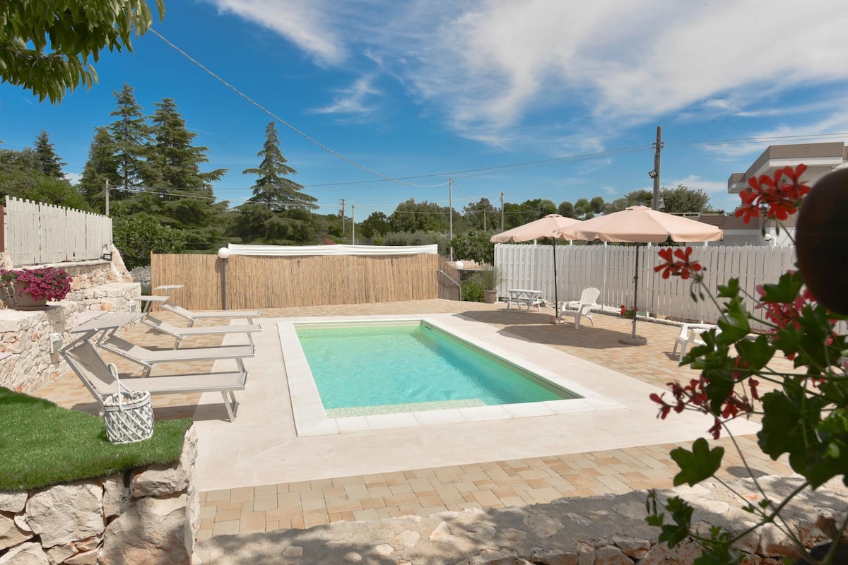 A private pool area is shown, featuring a clear blue water pool surrounded by a stone patio. Sun loungers are arranged around the pool, and shaded seating under umbrellas is visible. Lush greenery and flowering plants enhance the outdoor space, providing a tranquil atmosphere.