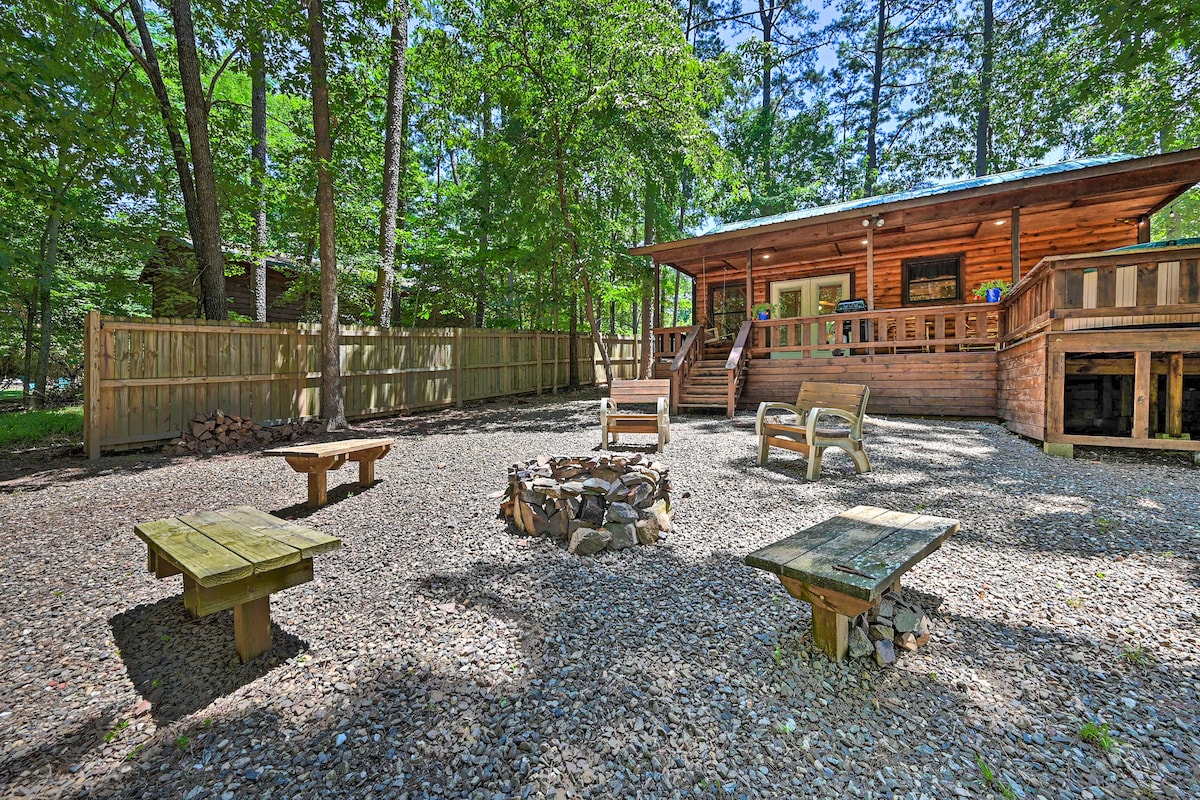 An outdoor space features a gravel yard with a fire pit surrounded by wooden benches and picnic tables. The cabin, with wooden exterior and a covered deck, is visible in the background, partially shaded by surrounding trees.