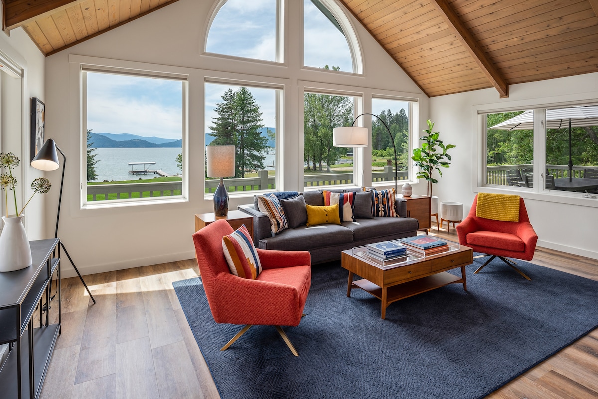 A bright living room features large windows that provide stunning views of the waterfront and surrounding landscape. Contemporary furnishings include a comfortable gray sofa and two vibrant red chairs, complemented by a wooden coffee table and area rug. Natural light fills the space, creating an open and airy atmosphere.