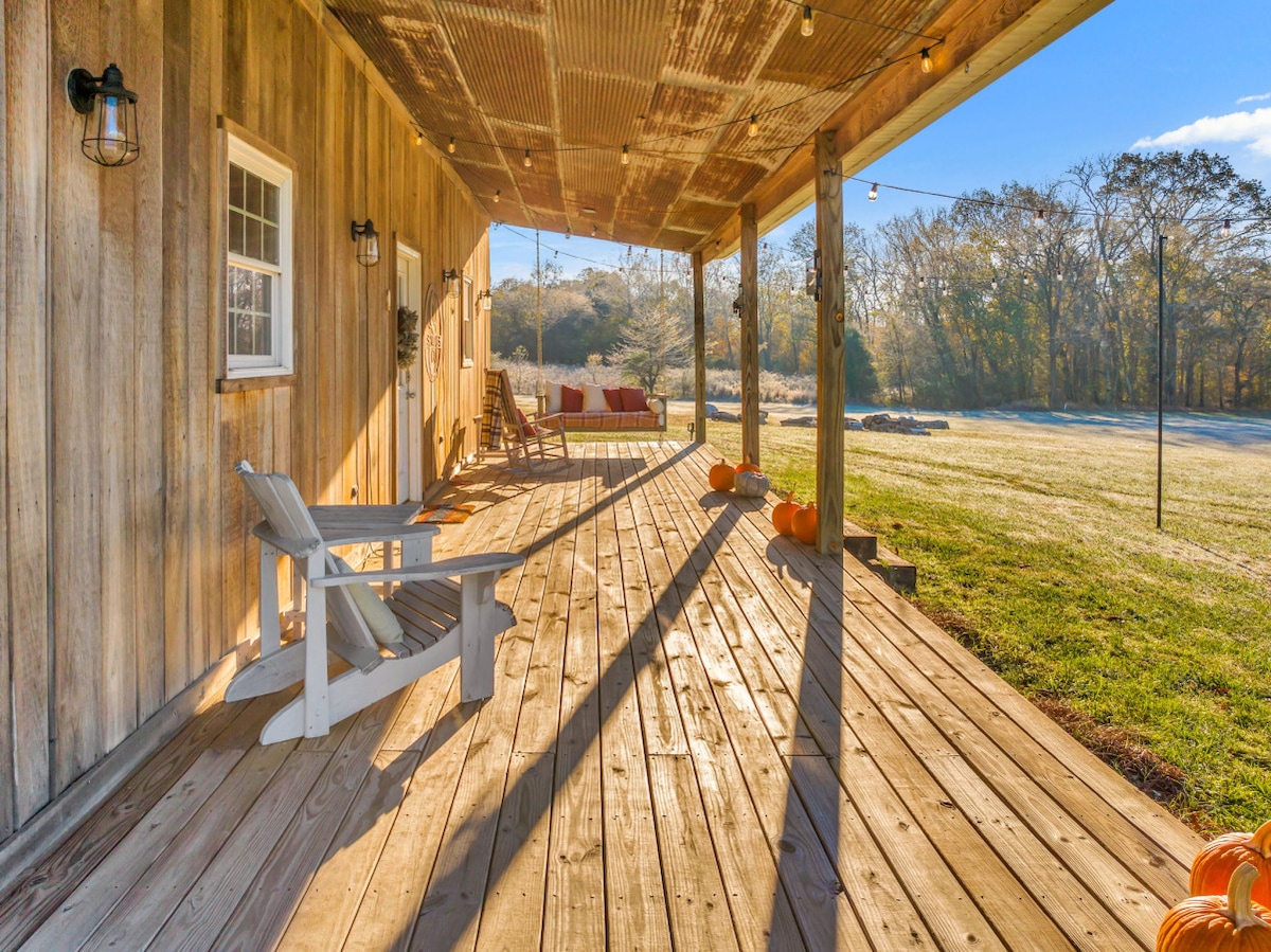 A spacious wooden porch is showcased, featuring an inviting swing and rocking chair. Soft sunlight casts shadows across the planked flooring, while pumpkins are scattered along the edges. A lush green lawn and trees are visible in the background, enhancing the serene atmosphere.