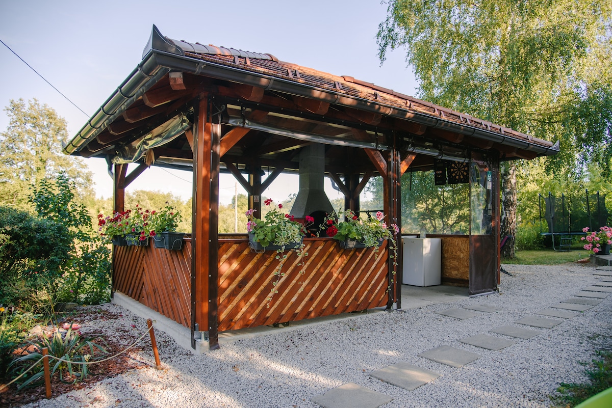 A rustic outdoor grill area is enclosed by wooden beams and adorned with blooming flowers. Gravel pathways lead to the structure, creating a charming environment. The open design allows for comfortable gatherings, while tall trees provide greenery in the background.