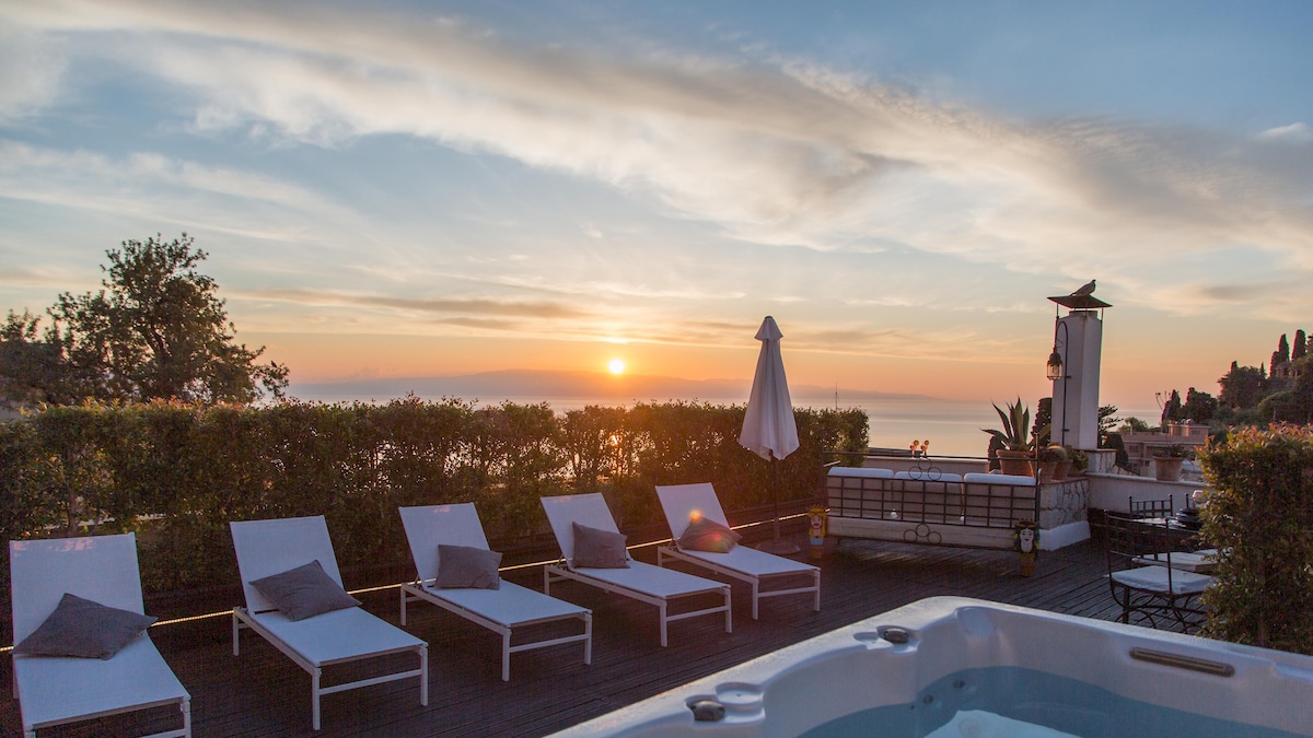 A panoramic terrace is shown at sunset, featuring several loungers and a jacuzzi beside lush greenery. The horizon showcases a vibrant sky with colors transitioning from orange to blue, creating a serene atmosphere for relaxation.