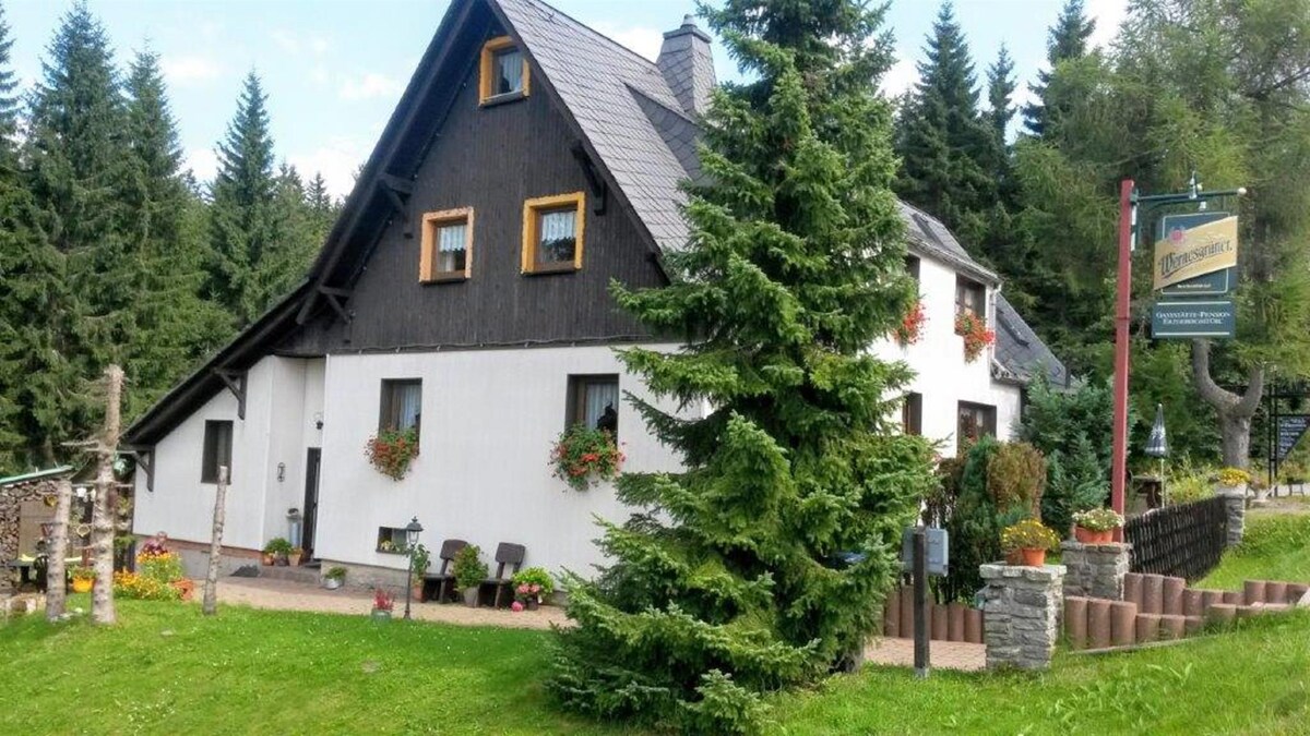 A charming two-story house is surrounded by lush greenery and a variety of flowering plants. The building features a combination of dark wooden and white siding, with flower boxes beneath the windows. A pathway leads to the entrance, complemented by neatly arranged stones.