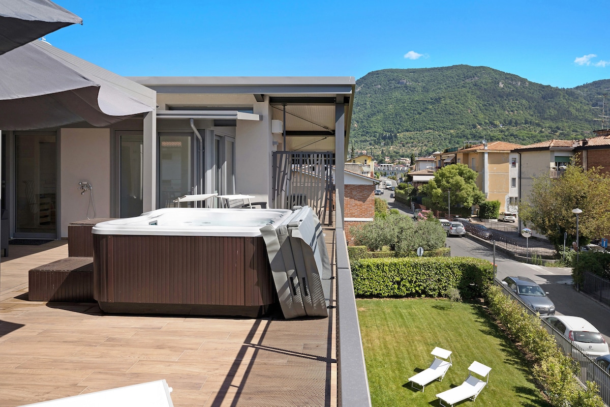 An outdoor terrace offers seating options with sun loungers and a hot tub, surrounded by greenery. A view of the nearby mountains and clear blue sky enhances the tranquil setting, while the wooden deck provides a warm, inviting space for relaxation.