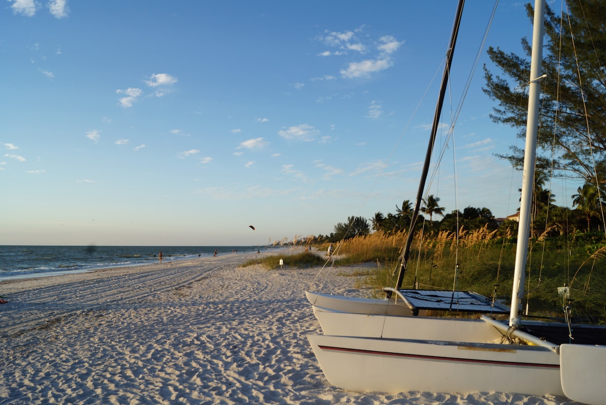A serene beach scene is depicted with two sailboats positioned on the sandy shore. In the background, gentle waves meet the coastline, while distant figures can be seen strolling along the water's edge. Lush greenery and palm trees frame the picturesque setting under a clear blue sky.