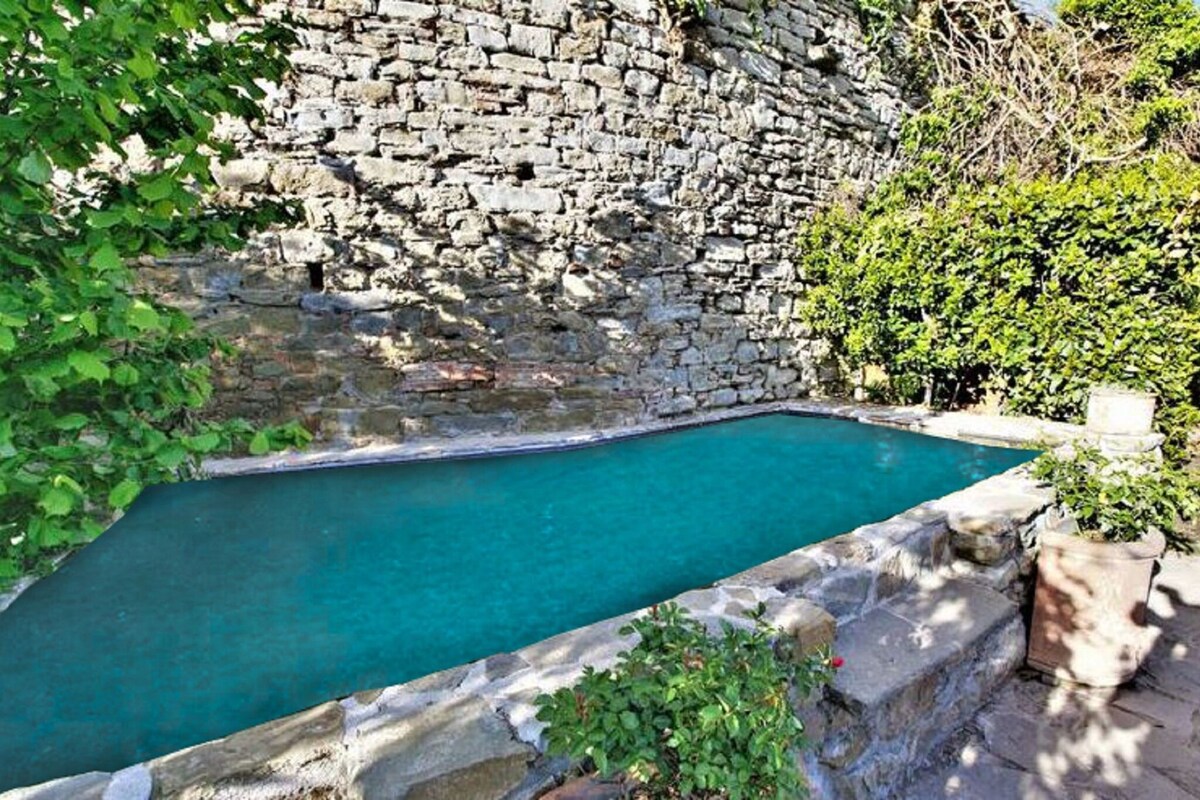 An outdoor swimming pool is framed by stone walls and lush green foliage. The pool's water is a serene turquoise, inviting relaxation. Surrounding the pool are potted plants and natural vegetation, creating a tranquil atmosphere.
