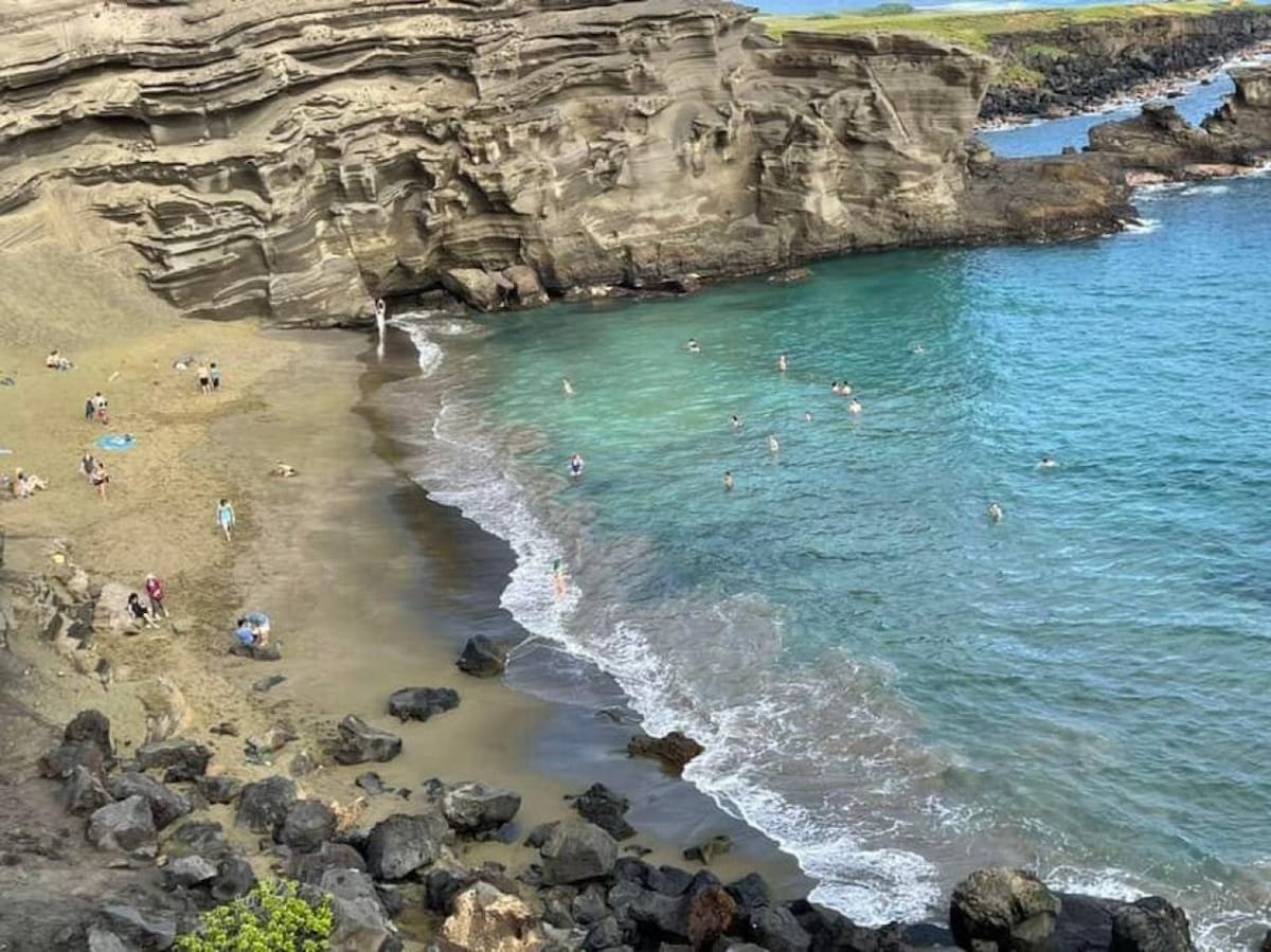 A view of Papakōlea Beach features striking green sand and rugged cliffs. Visitors can be seen enjoying the water and sunbathing on the sandy shore. Dark volcanic rocks line the beach, contrasting with the clear blue water, creating a captivating coastal landscape.