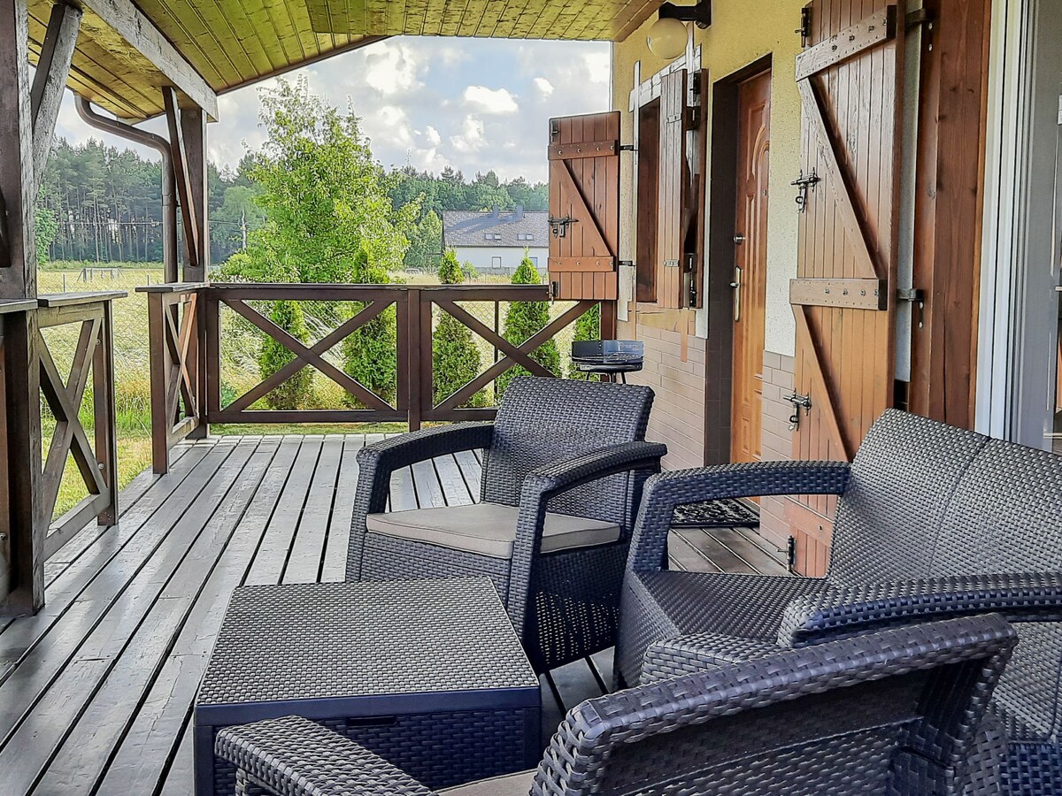 A spacious terrace features two rattan chairs and a small table, overlooking a green countryside view. The wooden structure is partially covered, with railings that complement the rustic style. Shutters and doors are visible, adding to the inviting atmosphere of the outdoor space.