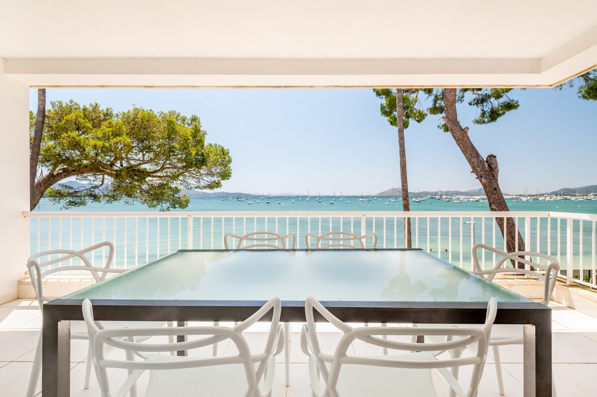 A spacious outdoor terrace features a large glass table surrounded by six white chairs. Clear views of the sea and boats in the distance are framed by two tall trees, creating a relaxing environment for enjoying the coastal scenery.