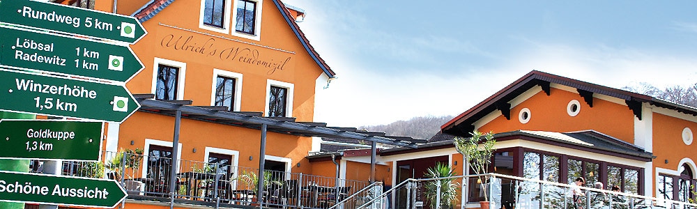 The exterior of a vibrant orange building featuring a modern architectural design is visible. A covered terrace area provides outdoor seating options. Directional signs indicate nearby attractions, emphasizing the establishment's proximity to various scenic locations.