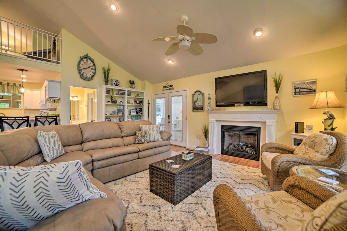 A cozy living area features a neutral sectional sofa and two patterned armchairs, arranged around a textured coffee table. A gas fireplace is situated beneath a large flat-screen TV. Bright yellow walls and well-placed decor enhance the inviting atmosphere, with natural light streaming in through multiple windows.