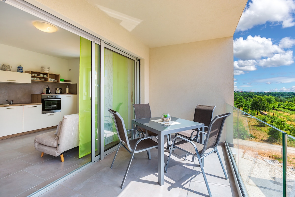 A spacious balcony is shown, featuring a dining table surrounded by four chairs. A modern kitchenette is visible through the glass doors, with the picturesque landscape of greenery stretching into the distance. Natural light floods the area, enhancing the inviting atmosphere.