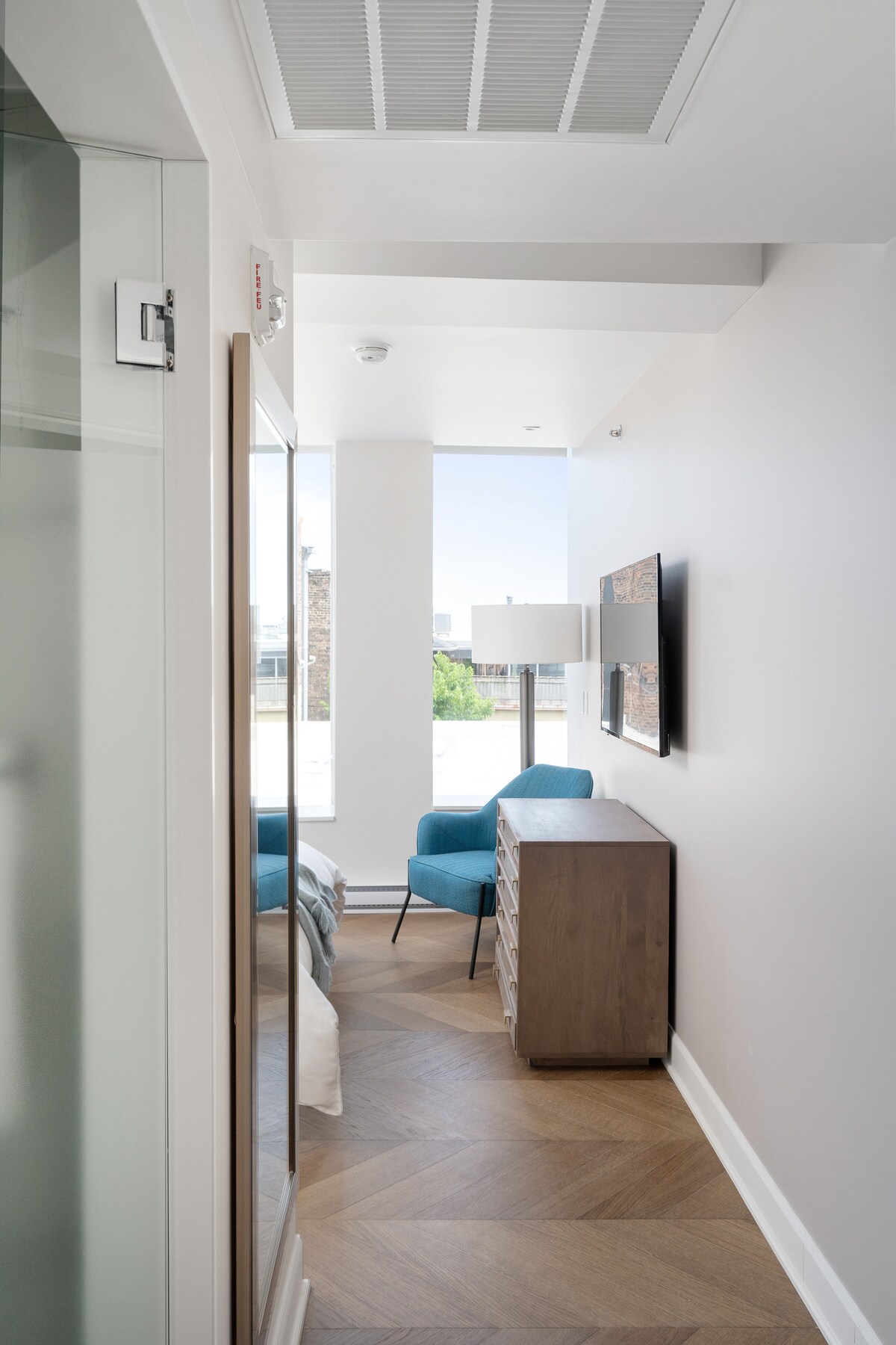 A narrow hallway features a combination of light wood and sleek surfaces. To the left, glass doors lead to a shower area, while a modern chest of drawers stands nearby. A comfortable blue chair is positioned next to a wall-mounted television, with soft natural light illuminating the space.
