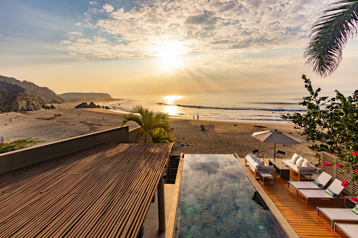 An expansive view showcases a beachfront infinity pool overlooking the ocean, with the sun rising above the horizon. Soft waves lap against the shore, while plush loungers are positioned along the pool deck. Lush greenery frames the scene, blending natural beauty with modern design.