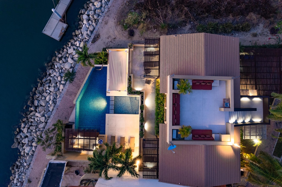An aerial view of the villa reveals a well-designed layout with a private infinity pool and swim-up bar, surrounded by tropical landscaping. The rooftop terrace is visible, showcasing outdoor seating under a shaded area. The villa is positioned next to a calm waterfront.