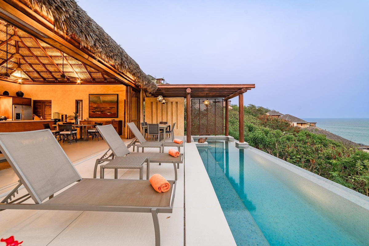A serene outdoor space features a sleek infinity pool surrounded by lounge chairs, each adorned with soft orange towels. Lush greenery frames the background, leading to expansive ocean views under a soft evening sky.