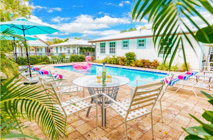 A shared pool area is framed by lush green foliage, featuring a circular table with four metal chairs positioned nearby. Colorful lounge chairs line the poolside, with vibrant beach towels draping over them. A pastel pink inflatable float rests in the inviting pool under a clear blue sky.