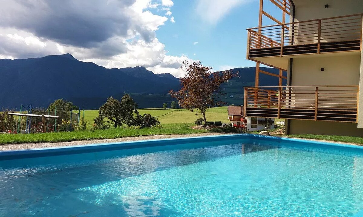 A shared outdoor pool is visible, set against a backdrop of distant mountains and open fields. The water reflects the sky, while wooden balconies are seen above, providing an inviting space for relaxation.